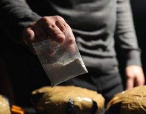 Man's hand holds a small plastic bag with white powder, with larger packages wrapped in brown tape in the background.
