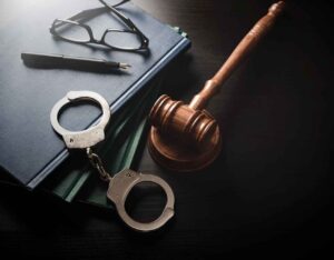 A top-down view of a gavel, handcuffs, a pair of glasses, and notebooks on a dark surface, symbolizing law and justice.