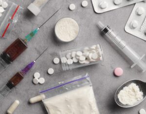 Various drug-related items are shown on a gray surface, including syringes, pills, and white powder.